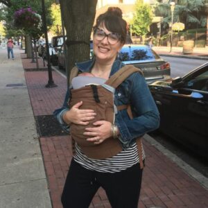 Heather is pictured on a sidewalk with her hair up in a bun, bangs, large cat-eye glasses, a jean jacket, a black and white striped shirt and black pants. She is holding a baby in a baby carrier across her chest.