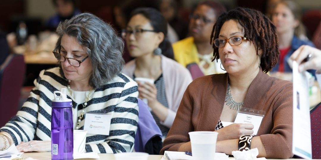 Two people sitting next to each other at conference. One is looking down and the other is looking at the unseen presenter. Other attendees are in the background.