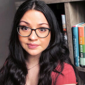Photo of Sandra with large black rimmed glasses on and long hair and a red shirt sitting in front of a bookcase