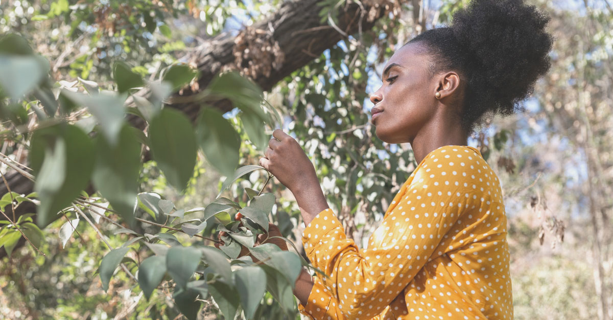 A Black woman with her hair up is wearing a long-sleeve yellow shirt. She is holding leaves and surrounded by greenery