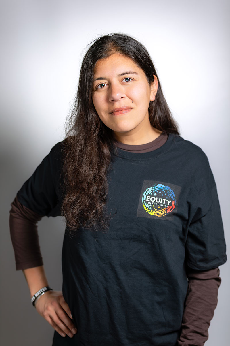 Niki Jagpal is pictured smiling at the camera with long black hair and her arm on her hip. She is wearing a black shirt with the Equity In The Center logo